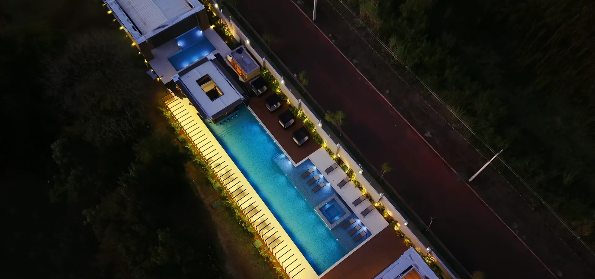 Vista aérea noturna do clube do Infinitus, com a piscina e o deck iluminados ao longo do clubhouse